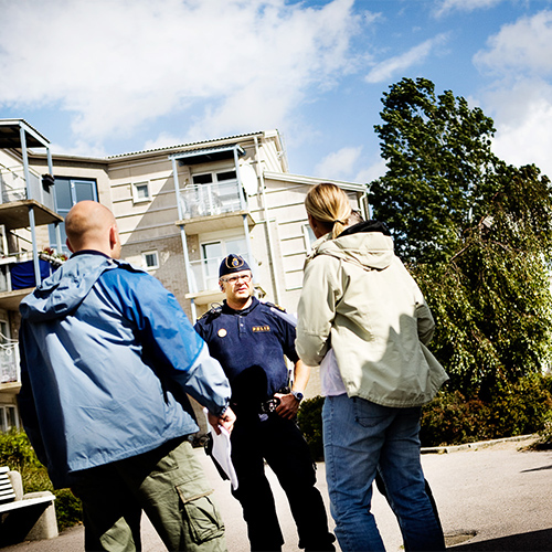 Polis i samtal med två personer utomhus framför ett bostadshus, fotograferad i ett redaktionellt sammanhang.