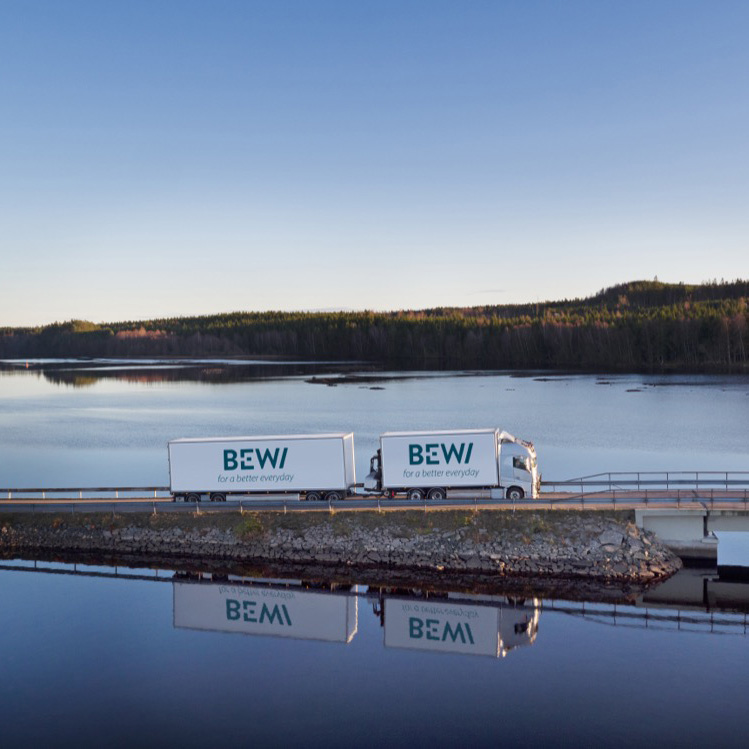 Drönarbild av en BEWI-lastbil på en bro, speglad i en stilla sjö med skog i bakgrunden.