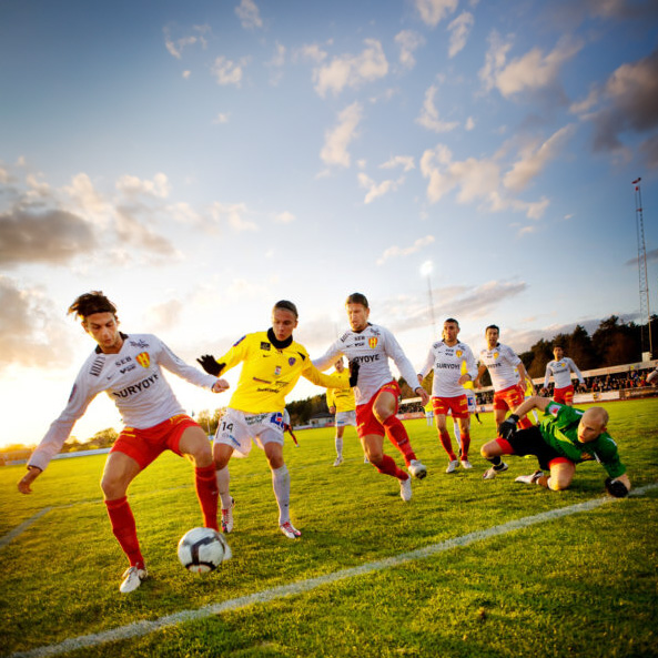 Intensiv fotbollsmatch fotograferad under en solnedgång, spelare kämpar om bollen nära mållinjen.